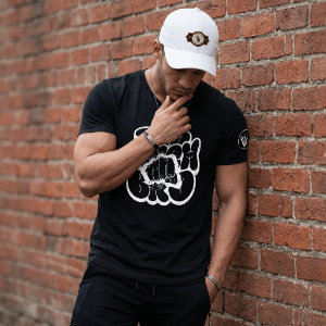 Model Wearing Black Too Much Bro T-shirt by Brick Wall Model wearing a black Too Much Bro T-shirt and white cap while standing beside a brick wall.