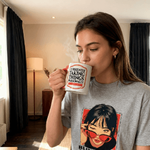 Woman holding Whuploo mug while wearing graphic T-shirt Woman holding a Currently Taking Things Personally mug while wearing a grey Blink Twice If It Grows T-shirt indoors.