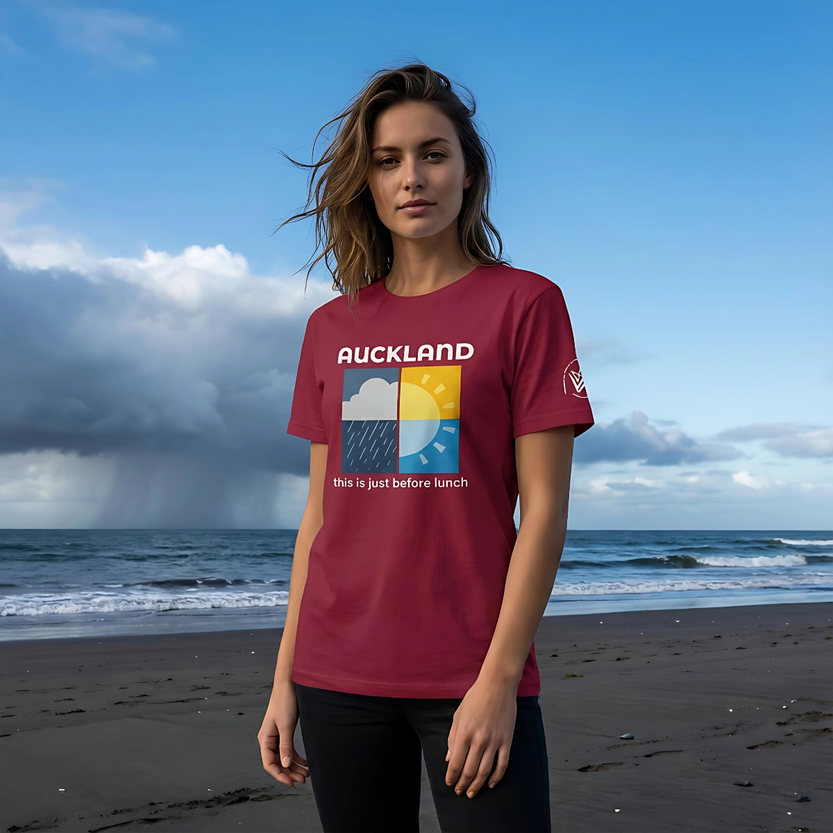 Woman wearing a cardinal red Auckland Weather T-shirt while standing on a beach with cloudy weather in the background