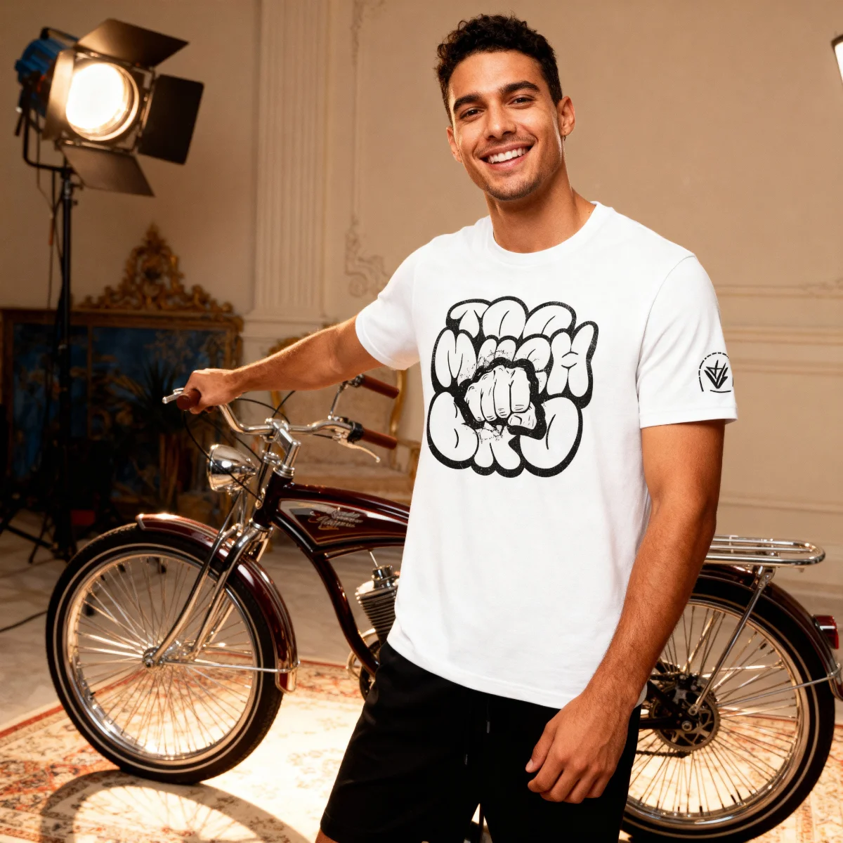Model wearing a white Too Much Bro graphic T-shirt with a sleeve logo, standing indoors beside a bicycle.