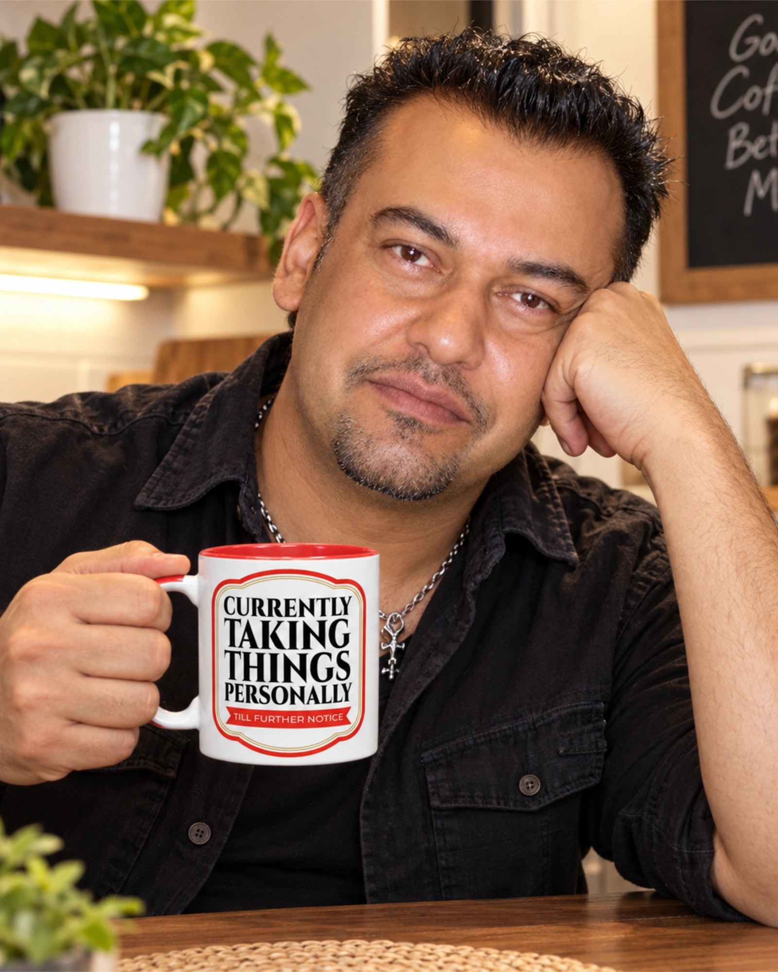 Man resting his head on his hand while holding a Currently Taking Things Personally coffee mug at a kitchen table.