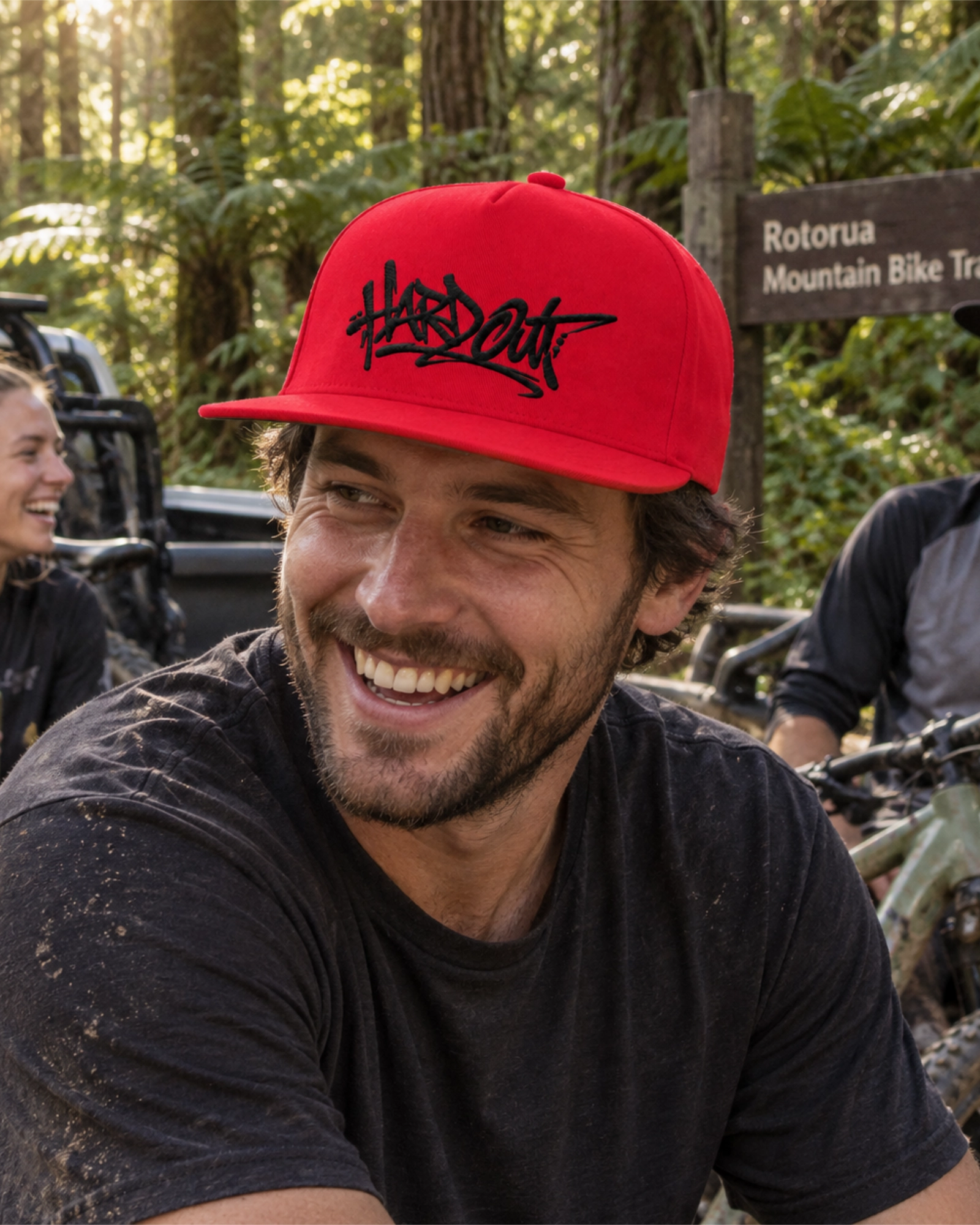 Man wearing a red HARDOUT flat bill snapback cap with black embroidery at a mountain bike trailhead