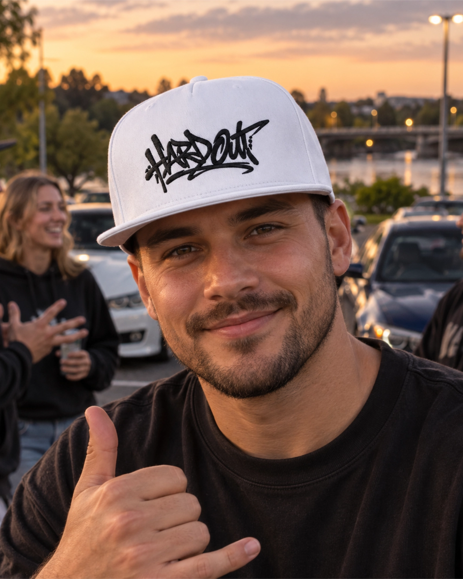 Man wearing a white HARDOUT flat bill snapback cap with black embroidery at an evening car meet