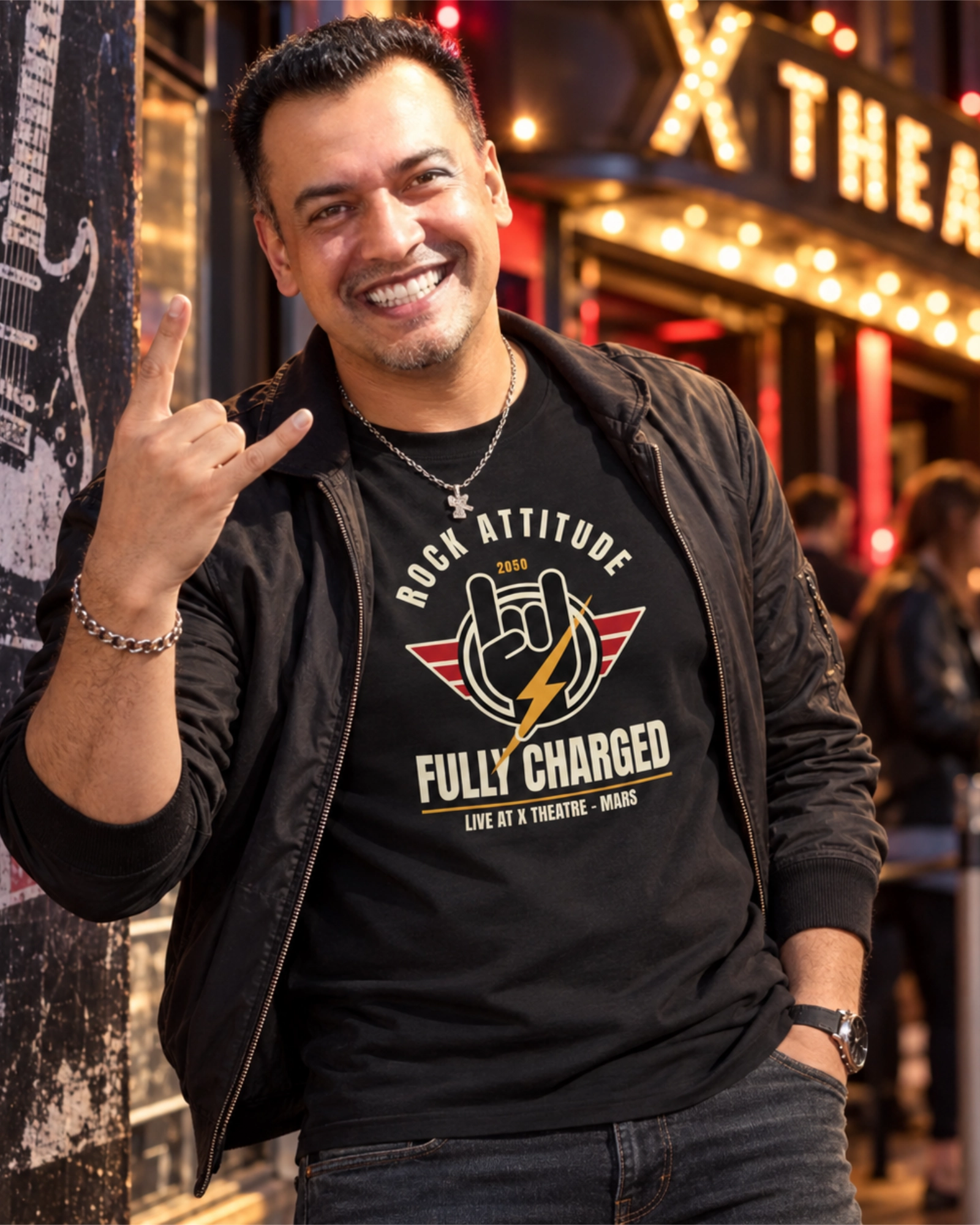 Smiling man making the rock horns gesture outside an illuminated music venue, wearing a black Rock Attitude Fully Charged graphic T-shirt.
