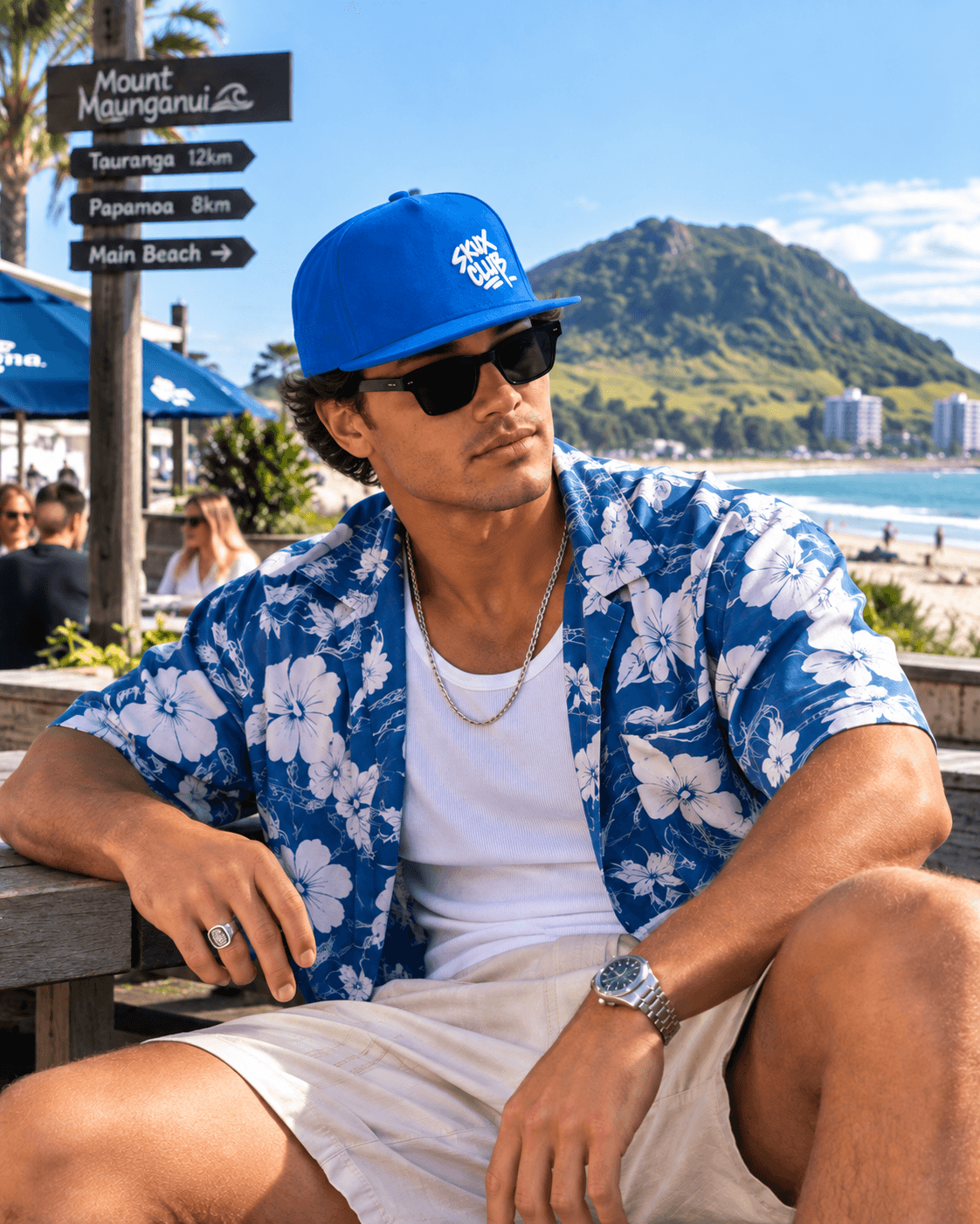 Man wearing a royal blue SKUX CLUB flat bill cap with white handstyle graffiti embroidery, black sunglasses and a blue floral shirt at Mount Maunganui.