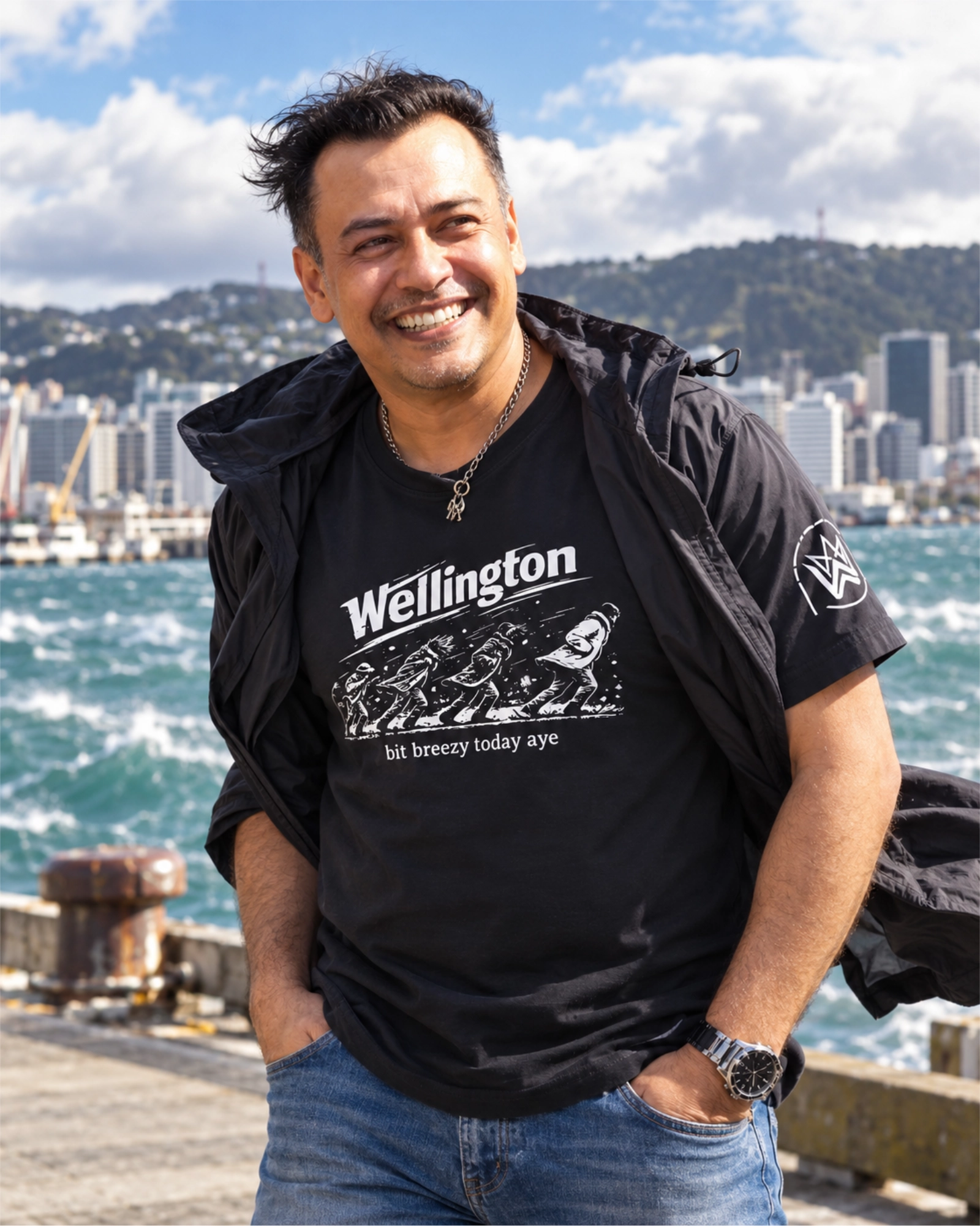 Smiling man wearing a black Wellington “bit breezy today aye” T-shirt, with his jacket blowing in the wind beside Wellington Harbour.